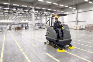 Cleaner on a warehouse concrete floor beside racking and a roller door